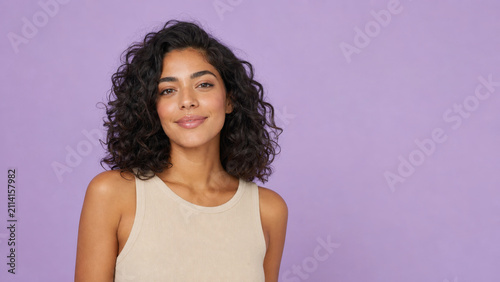 Confident woman with curly dark hair wearing beige sleeveless top posing against pastel lavender background with gentle smile and relaxed