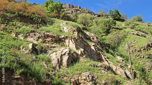Sun-lit sandstone rock formations emerge through wild grass on a terraced hillside, leading up to traditional stone houses and shrubs beneath a clear blue sky.
📍 Hawraman Takht, Kurdistan, Iran 