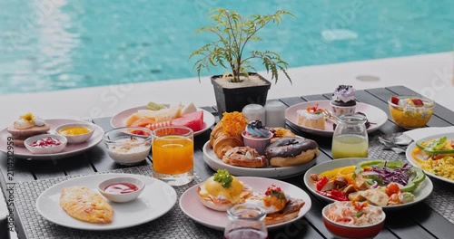 Breakfast dishes including eggs benedict, croissants, and fresh fruit juice set on table beside swimming pool, luxury resort dining, morning meal at vacation hotel