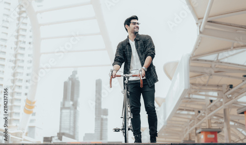 Foto Urban Commuter: Young man with beard stands with his vintage bicycle in front of modern buildings, enjoying a moment of reflection amidst the city bustle