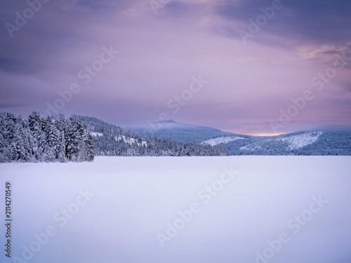 Peaceful winter landscape with a frozen snow-covered lake surrounded by frost-covered forest and rolling hills beneath a soft pink and purple sunset sky in Swedish Lapland.