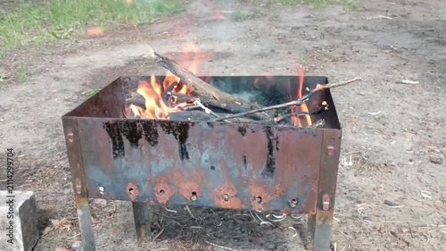 Flames erupt from a decorative metal grill set on the ground and grass. Burning wood glows within, smoke rising.