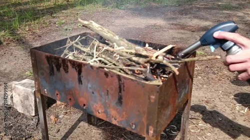 A hand lights kindling in a metal brazier with a blue-black gas torch. Thin branches and twigs lie in the brazier on a patch of grassy ground.