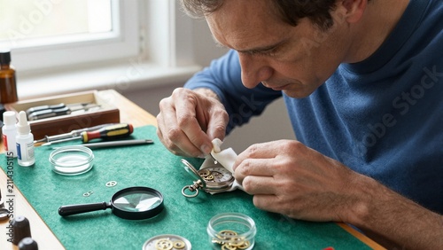 Skilled artisan meticulously cleans an antique silver pocket watch using specialized tools and a soft cloth on a green work surface.