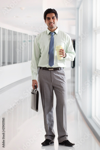 Businessman with bag and coffee cup standing in office corridor 