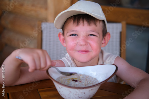 Little boy wearing baseball cap eating oatmeal from white bowl. Child sitting at table and enjoying healthy breakfast. Smiling kid having meal at home. Morning nutrition concept.