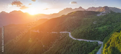 Panel kuchenny z motywem Winding mountain road traversing a lush green forest with dramatic sun flare and mountain peaks during a vibrant sunset