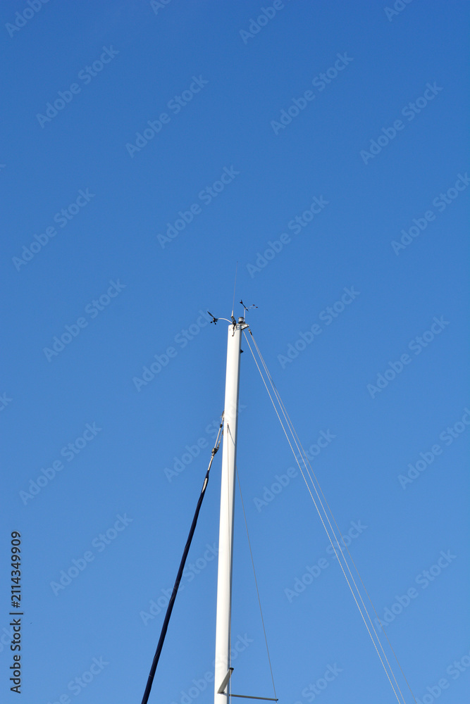 Fototapeta premium The white masthead of a sailboat features a wind vane anemometer and radio antenna against a solid blue sky background