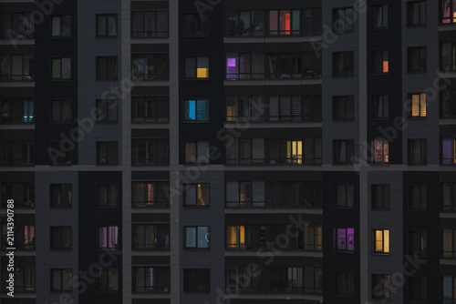 Modern apartment building facade at dusk with illuminated windows in different colors. Urban residential architecture creating a geometric pattern of light, shadow and everyday city life.
