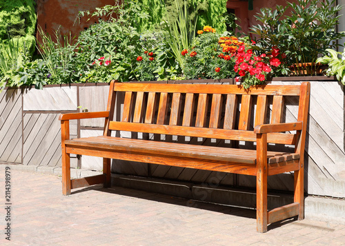 Wooden park bench garden photo. Empty varnished timber seat stands on brick pavement in front of blooming flowerbed with red orange blossoms. Classical outdoor backyard patio furniture design closeup 