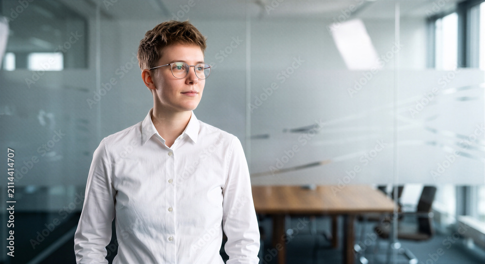 Obraz premium Young business professional standing in a modern glass office, wearing glasses and a white shirt, looking away with a confident expression