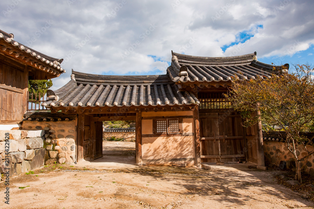 Fototapeta premium Exterior View of Traditional Korean Hanok Entrance Gate Featuring Courtyard, Stone Wall, and Tiled Roofs Under Cloudy Sky