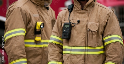 Two firefighters in tan protective suits with yellow reflective stripes stand ready