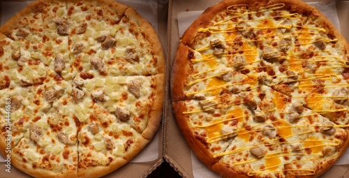 Two round pizzas in cardboard boxes. On the left is a classic Hawaiian pizza with chicken and pineapple pieces on mozzarella cheese. On the right is a cheese pizza with chicken, drizzled with sause