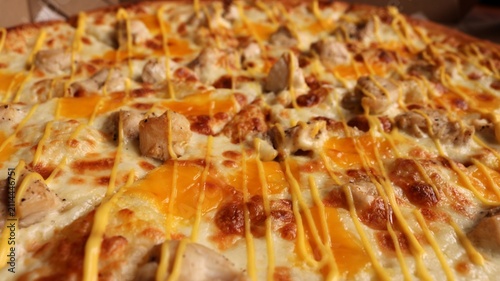 A close-up of a delicious pizza with chicken breast pieces topped with melted cheese. The dish is topped with stripes of yellow sauce, and delicious islands of cheddar are baked into the fluffy crust.