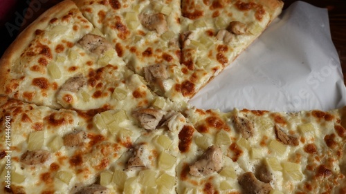 A delicious Hawaiian pizza with chicken and pineapple. It's cut into slices, with one piece missing. The crispy mozzarella crust and chunks of sirloin are visible. The dish sits on white baking paper.