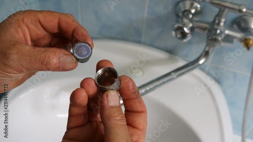 A man's hands hold a disassembled faucet aerator: a round mesh filter and a metal sleeve attachment. The process takes place over a white sink in a bathroom with blue tile on the wall.