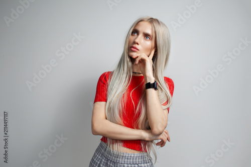 Young pretty blonde girl in red shirt looking thoughtfully away isolated on grey background.