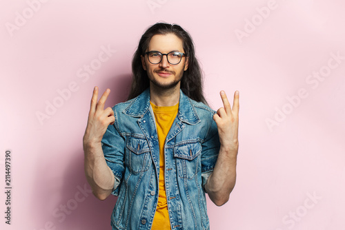 Young confident man with long hair showing peace gesture isolated on pastel pink background.