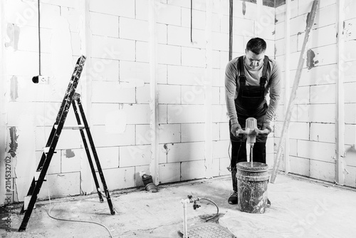 A male plasterer in overalls mixes gypsum mortar for plastering walls in a bucket using an electric mixer black and white photo