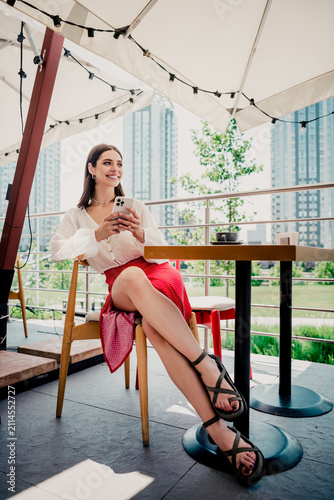 фотографія Young woman sitting at an outdoor cafe terrace in the city, smiling and using he