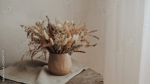 A beautiful bouquet of dried wild flowers in a rustic clay ceramic vase sitting on a soft linen cloth on an old wooden table in a cozy home interior with bright natural soft light