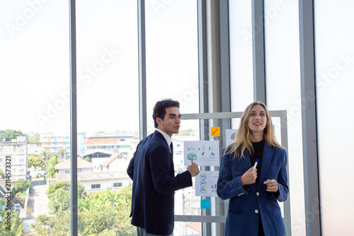Two business professionals discussing marketing strategy and business analytics in front of a glass board with charts and sticky notes.Corporate colleagues collaborating on project planning,