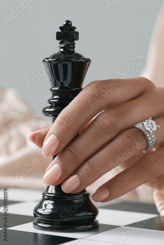 A jeweled hand grips a glossy black chess king on a board, symbolizing elegance, power, strategy and commitment.