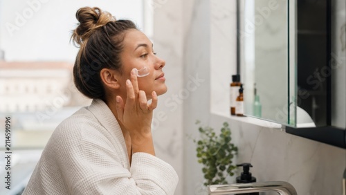 Young woman applying moisturizer in her morning bathroom routine captures a wellness moment with natural white light and skincare