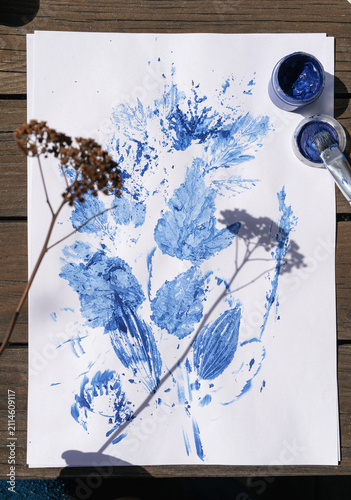 Картината върху платно Dry plant stem with seeds casting a shadow on a white background with blue painted leaf and flower imprints
