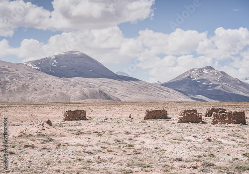 Ruins of ancient Pamir dwellings in the Tien Shan Mountains of Tajikistan on a high plateau with a mountain with cliffs and snow in the background. Landscape in sunny weather