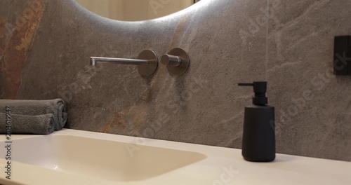 Backlit round mirror and wall mounted faucet above an integrated sink, camera glides left to right across dark stone cladding pale countertop folded towels and matte accessories