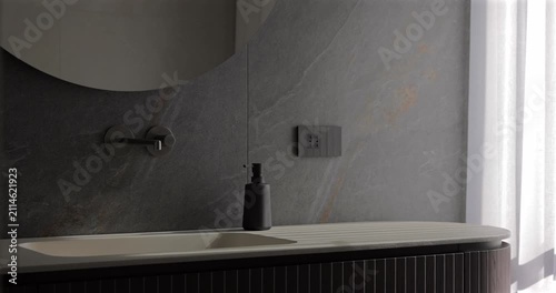 Bathroom interior with rounded mirror ribbed vanity and natural stone wall, static video in soft daylight as sheer white curtains move lightly by the window and gentle shadows settle across sink