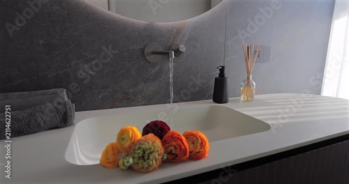 Bathroom sink with steady water flow over fresh flowers, static video close view of integrated basin wall mounted faucet dark stone wall folded towels reed diffuser