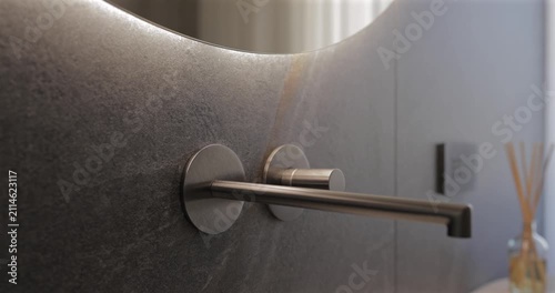Sensor activated mirror glow above a brushed wall faucet in dark stone bathroom, static close video follows hand passing beneath the rounded edge as soft backlight awakens reshapes premium sink area
