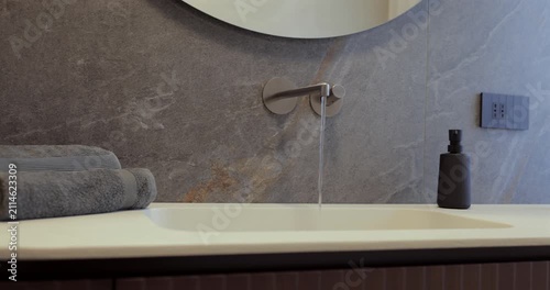 Bathroom interior with flowing water from wall faucet and curved wood vanity, left to right camera glide reveals the stone countertop round mirror folded towels black soap dispenser and reed diffuser