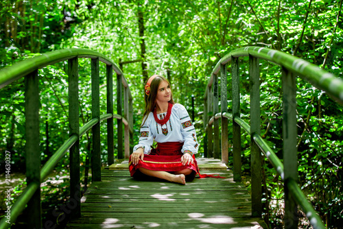 Young Ukrainian woman in traditional embroidered clothing crossing a forest bridge, symbolizing cultural roots, personal journey, continuity and transition.