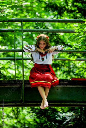 Young Ukrainian woman in traditional embroidered clothing crossing a forest bridge, symbolizing cultural roots, personal journey, continuity and transition.