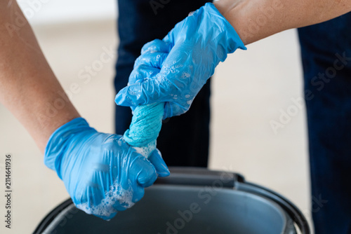 Close-up of hands in blue nitrile gloves wringing out a soapy microfiber cloth over a plastic bucket. Professional cleaning service or chores concept.