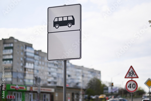 White rectangular road sign shows public transit stop. Standard navigation symbol with blank space marks passenger pickup point near buildings. Metal street sign guides city commuters.
