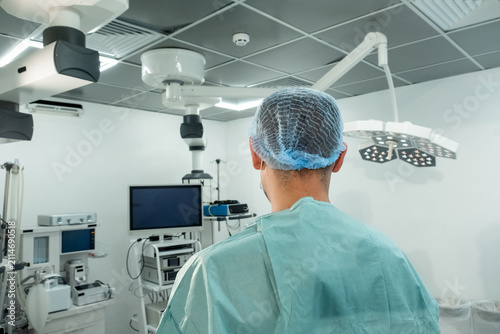 Man surgeon stands under overhead lights inside modern operating room. Focused medic checks surgical setup preparing room for procedure