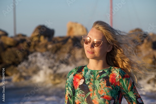 Bild auf Leinwand Blonde woman with windblown hair poses near a rocky shoreline with blurred sea spray behind her