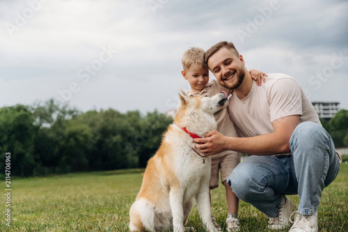 Caucasian father and son kneeling on grass petting a friendly Akita dog in a green park with trees and a cloudy sky in the background