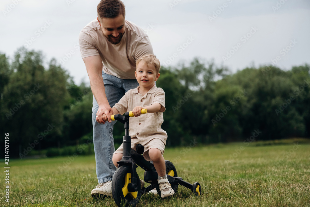 Obraz premium Father assists young boy riding a tricycle on grassy field, surrounded by trees and open space, showcasing a playful outdoor moment in a natural setting