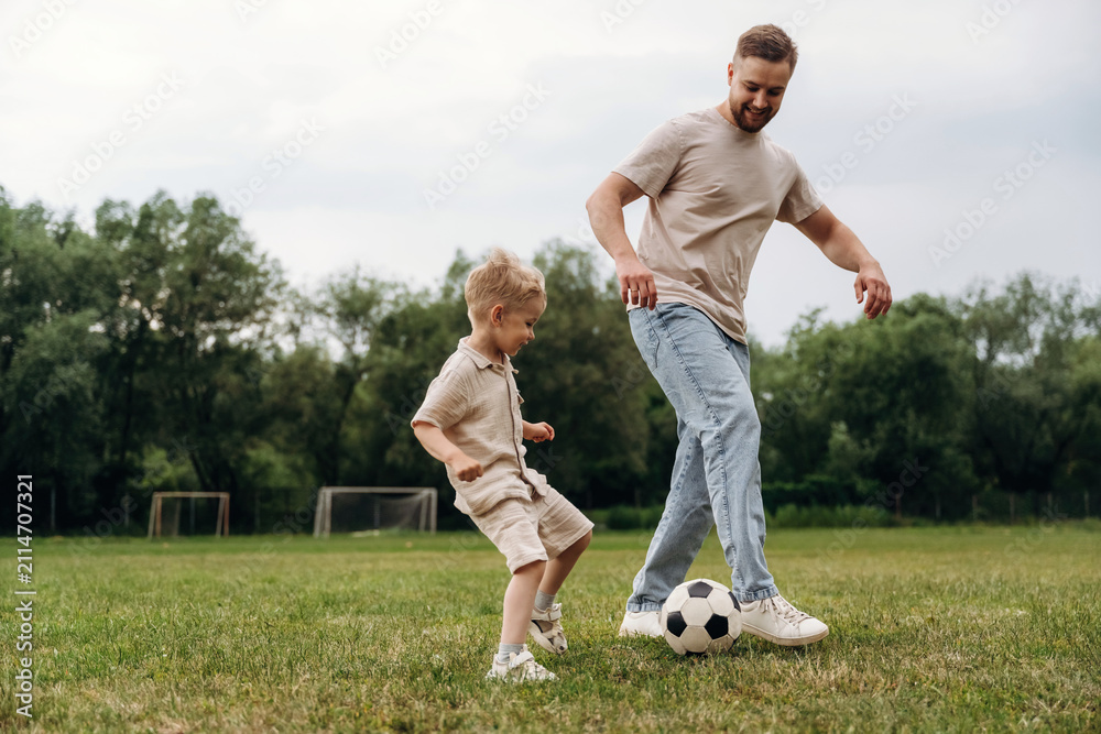 Obraz premium Father and son playing soccer on grassy field, child in beige outfit and father in casual wear, soccer ball at their feet, goalposts visible in the background