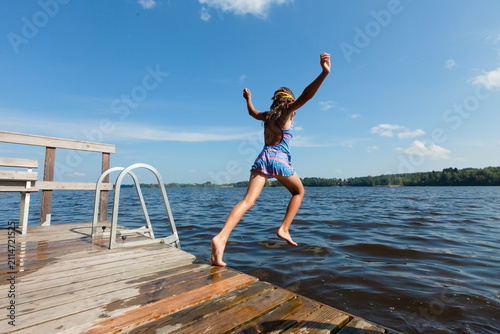 Family vacation joy as child jumps off wooden dock into lake on summer day. Little girl spends quality time outdoors with loved ones enjoying nature and water fun. Kid creates happy memories during.