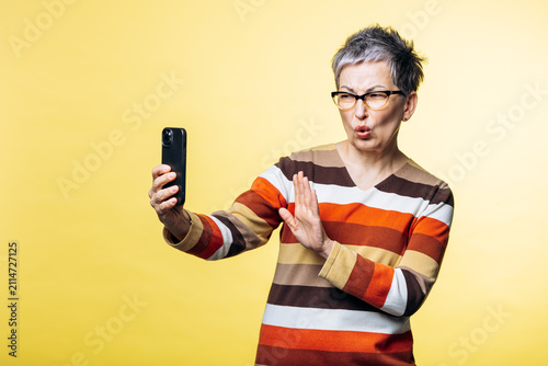 Bild auf Leinwand Senior woman posing with a funny face, holding a smartphone and taking a selfie