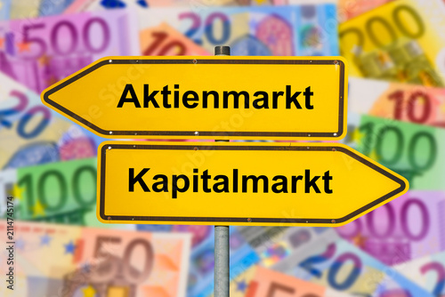 Zwei Wegweiser über einer Menge Euro-Bargeld zeigen in unterschiedliche Richtungen und sind beschriftet mit Aktienmarkt und Kapitalmarkt - FOTOMONTAGE