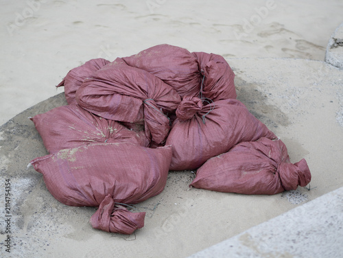 A pile of stacked purple-red woven plastic sacks filled with sand, tied at the ends and placed on a concrete surface near sand.