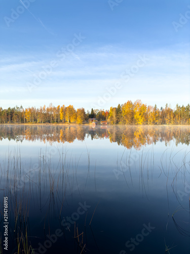 Fotografie A serene view of a calm lake in Sweden during autumn, featuring vibrant fall foliage reflecting on the mirror-like water and traditional red houses
